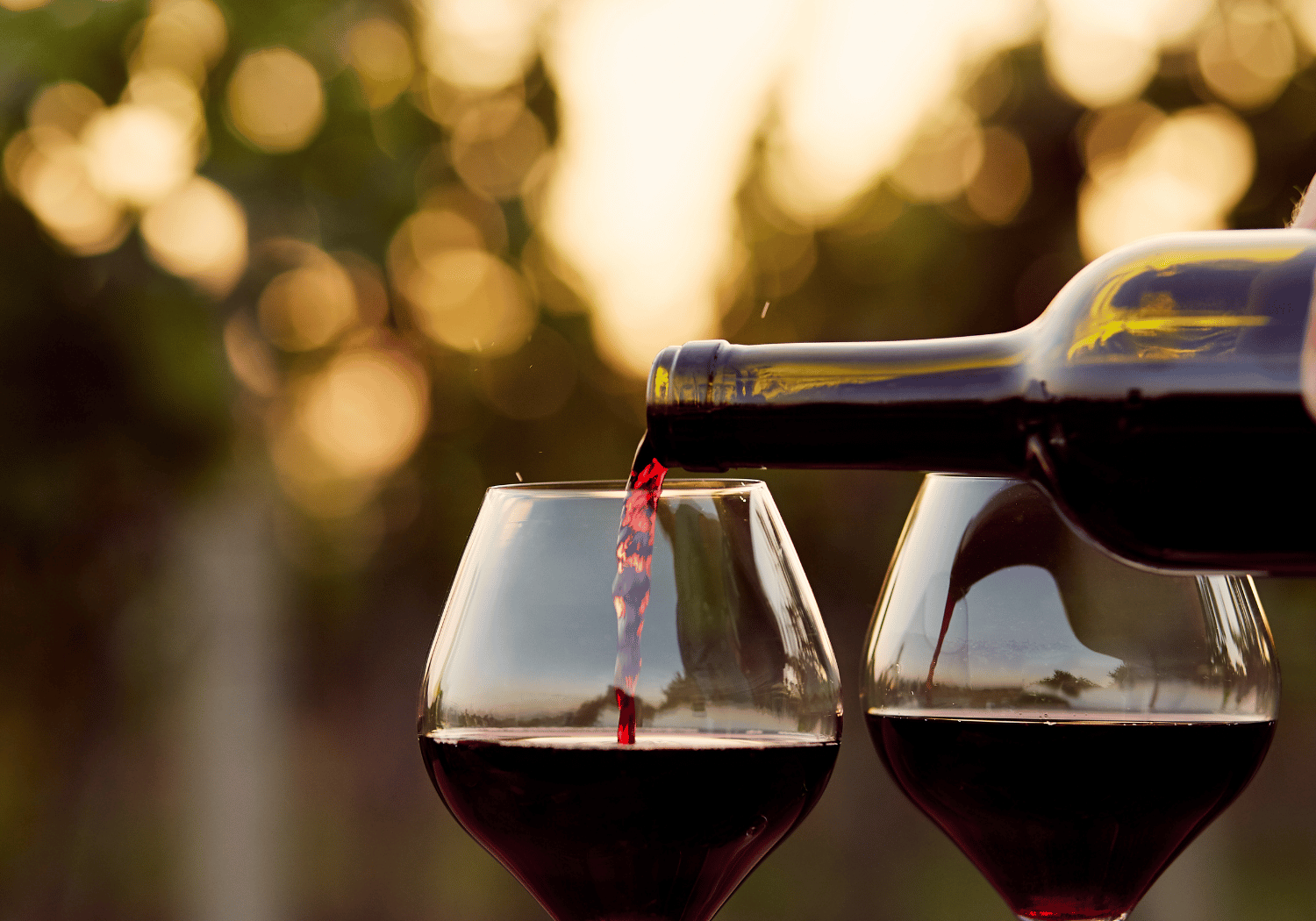 Red wine being poured from a bottle into two wine glasses outdoors with warm sunset bokeh in the background.